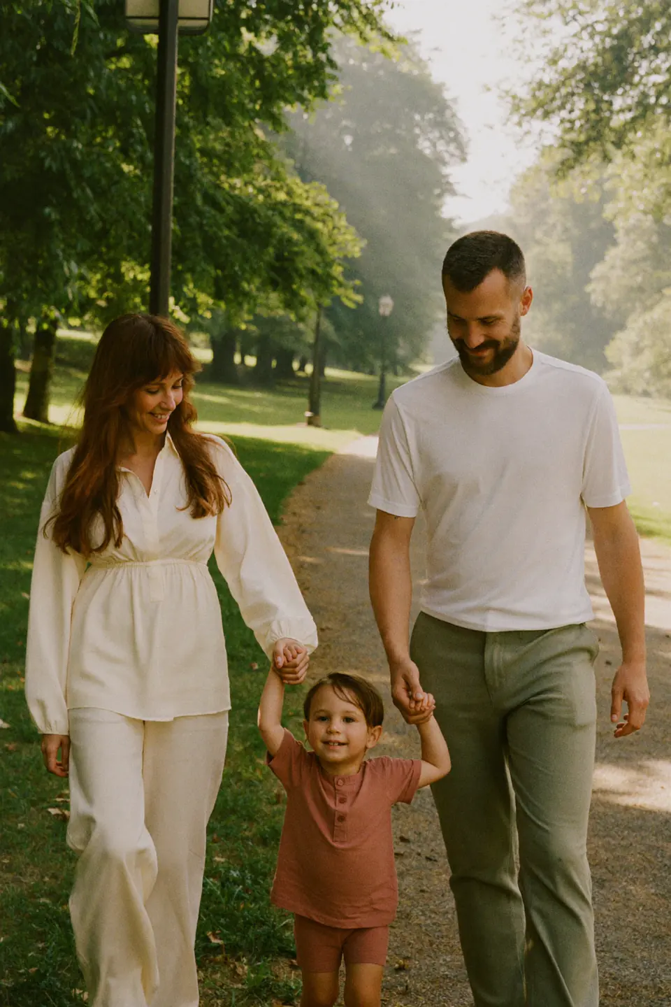 A mother, father and their young child walking hand in hand along a sunlit park path