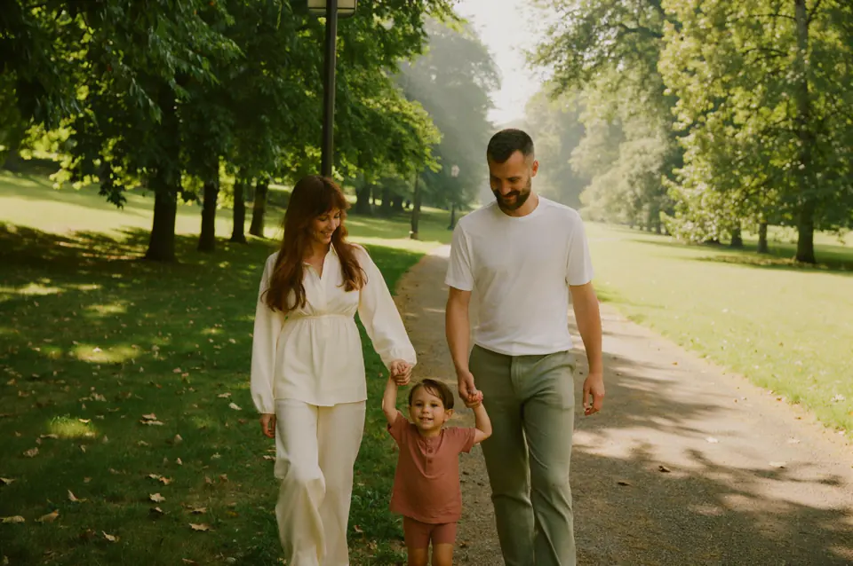 A mother, father and their young child walking hand in hand along a sunlit park path